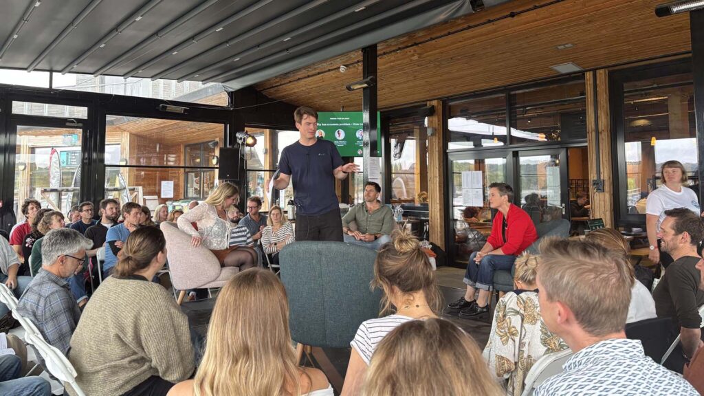 A man stands in the center of a conference room, speaking to people sat on chairs around him